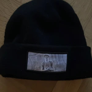 Svart mössa med tryckt patch - Svart mössa med uppvikt kant och en rektangulär patch framtill med tryckt motiv av en person och graffiti-liknande bakgrund. Mössan har enkel, klassisk form och passar dig som gillar streetstyle. Materialet känns mjukt och stretchigt, troligen bomull eller polyester.