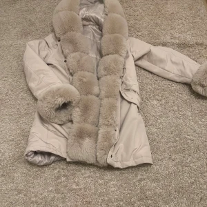 Beige pälsjacka med fluffig krage - Säljer en beige pälsjacka med superfluffig krage och muddar i ljus päls. Jackan har dragkedja, snörning i midjan och en ficka med dragkedja. Insidan är quiltad och jackan är perfekt för kalla dagar. Stilren och lyxig vibe.