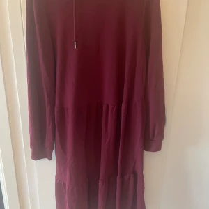 Vinröd långklänning från Jacqueline de Yong - Vinröd långklänning från Jacqueline de Yong med huva och långa ärmar. Klänningen har en avslappnad passform och är tillverkad i mjuk blandning av polyester och viskos. Snygg volangdetalj nedtill och enkel design utan mönster.