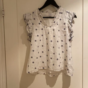 Vit blommig blus med volang från Lindex - Supersöt vit blus från Lindex med små blå blommor och volangdetaljer på ärmarna. Blusen har v-ringning och är ärmlös med svarta vågiga kanter på volangerna. Perfekt för en fräsch och somrig look!