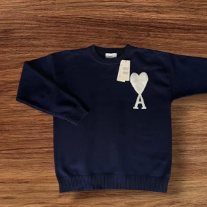 Mörkblå stickad tröja från Ami - Snygg mörkblå stickad tröja från Ami med vit hjärtlogga och bokstaven A på bröstet. Rund hals och ribbade muddar vid ärmslut och nederkant. Perfekt för dig som gillar stilrena detaljer och vill ha något som sticker ut lite extra.