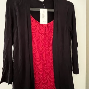 Svart långärmad blus Amalia S - Svart långärmad blus från Amalia i storlek S. Blusen  har en röd insida med dekorativt mönster framtill och en enkel, rak passform. Materialet känns mjukt och lätt, perfekt för dig som gillar stilrena plagg med en twist.