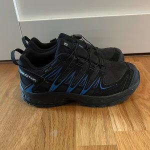 Svarta Salomon Xapro sneakers 37 - Salomon Xapro sneakers i svart med blå detaljer och grov sula för extra grepp. Skorna har snörning med snabbspänne och är tillverkade i slitstark syntet med meshpaneler för ventilation.  Använda 1 gång