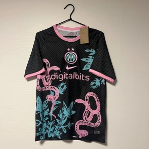 Inter tröja rosa orm - Inter fotbollströja från Nike i svart med rosa och turkosa detaljer. Storlek M. Snyggt ormmotiv och växtgrafik framtill, rosa logga och detaljer vid hals och ärmslut. Tillverkad i lätt och ventilerande polyester, perfekt för match eller träning.