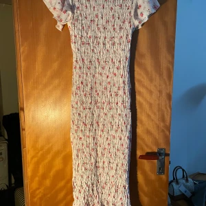 Vit långklänning med blommigt mönster - Supersöt långklänning i vit med små rosa blommor och korta puffärmar. Klänningen har smockad överdel och är figurnära med stretchigt material. Perfekt för sommarens alla tillfällen och ger en romantisk vibe med sitt blommiga mönster och mjuka tyg.