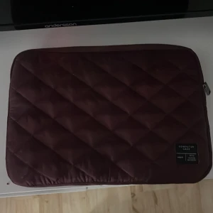 Quiltad vinröd datorväska Denim Makers - Quiltad datorväska i mörk vinröd färg från Denim Makers. Väskan har ett stilrent rutmönster och dragkedja på sidan. Perfekt för att skydda din laptop med snygg vibe och mjuk känsla. Märkt med 'Computer Case' på framsidan.