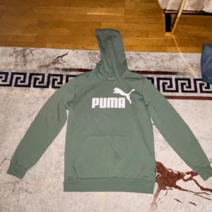 Grön hoodie från Puma med logga - Snygg grön hoodie från Puma med stor vit logga på bröstet. Tröjan har huva, långärmade ärmar och en praktisk magficka framtill. Perfekt för chill eller träning, och den har en klassisk, relaxed passform.