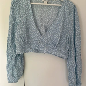 Blommig croppad blus från Monki - Söt ljusblå croppad blus från Monki med vitt blommönster. Blusen har v-ringning, långa puffiga ärmar och knappar framtill. Perfekt för dig som gillar romantiska och trendiga plagg med en avslappnad vibe.