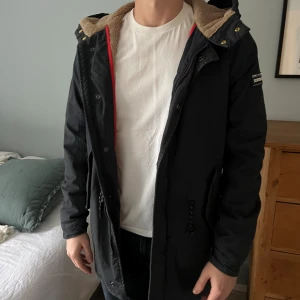 Mörkblå parkajacka från Scotch & Soda - Mörkblå parkajacka från Scotch & Soda med huva och beige teddyfoder. Jackan har snörning i midjan, dragkedja och tryckknappar framtill samt två stora fickor. Patch med logga på ärmen och annan snygg design. Perfekt för kyliga dagar. NYPRIS: Ca. 3500