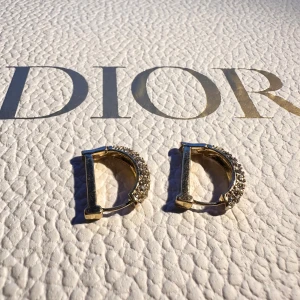 Dior guldörhängen med strass - Säljer ett par ikoniska örhängen från Dior i guldtonad metall, formade som bokstaven D. Ena sidan är täckt av glittrande strass som ger en lyxig och trendig vibe. Perfekta för dig som vill sticka ut med exklusiva accessoarer.box not included