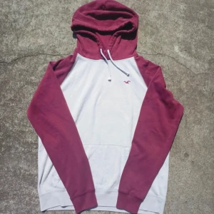 Hollister hoodie - Säljer en hoodie från Hollister i storlek M. Tröjan är vit med vinröda ärmar och huva, samt har en broderad Hollister-logga på bröstet. Klassisk känguruficka och snörning i huvan. Perfekt för dig som gillar en avslappnad stil.