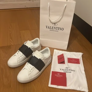 Valentino Garavani Open sneakers vit/svart - Snygga Valentino Garavani Open sneakers i vitt skinn med bred svart läderdetalj över vristen. Klassisk rund tå, vita snören och diskret logga på plösen. Kommer med originalpåse och certifikat. Perfekta för dig som gillar lyxiga och stilrena sneakers.