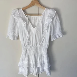 Vit kortklänning med volanger - Supersöt vit kortklänning med volanger och v-ringning. Klänningen har korta ärmar, smock i midjan och flera lager volanger nedtill. Tillverkad i lätt bomullstyg som ger en luftig känsla. Perfekt för sommarens alla tillfällen och ger en romantisk vibe.