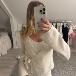 Vit omlottkofta med knyt - Supermysig vit omlottkofta med knyt i sidan och v-ringad hals. Koftan har långa ärmar och är gjord i ett mjukt stickat material som känns skönt mot huden. Perfekt att slänga på sig när det är lite kyligt ute. Enkel och stilren design som passar till allt.