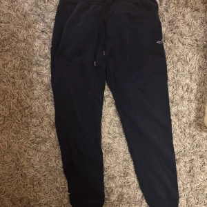 Mörkblå mjukisbyxor från Hollister - Mörkblå mjukisbyxor från Hollister med broderad logga på benet. Byxorna har dragsko i midjan och muddar vid bensluten för en skön och avslappnad passform. Perfekta för chill dagar eller när du vill vara extra bekväm.