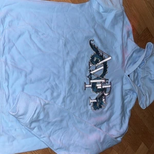 Ljusblå AMIRI hoodie med draktryck - Säljer en ljusblå hoodie från AMIRI med stort broderat drakmotiv och logga på bröstet. Tröjan har huva, magficka och långa ärmar. Perfekt för dig som gillar streetwear och vill sticka ut med snygga detaljer. Lite skrynklig bara
