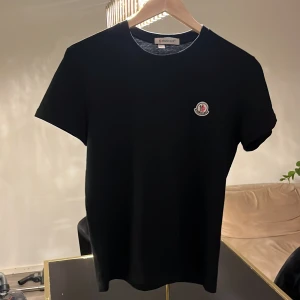 Svart Moncler t-shirt med logga - Svart t-shirt från Moncler i storlek S. Klassisk rund hals och korta ärmar. På bröstet sitter Monclers ikoniska broderade logga i vitt, rött och blått. T-shirten har en clean och stilren look, perfekt för dig som gillar exklusiva märken och enkel design.
