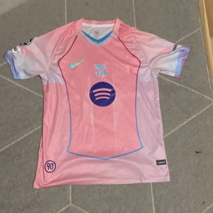 Barça Lamine Yamal rosa Nike tröja - Säljer en Barcelona fotbollströja med Lamine Yamal och nummer 10 på ryggen. Tröjan är rosa med lila och blå detaljer, har korta ärmar och är tillverkad i lätt polyester. Nike-logga och FCB-emblem på bröstet, samt Spotify och UNHCR tryck. UEFA Foundation-märke på ärmen.