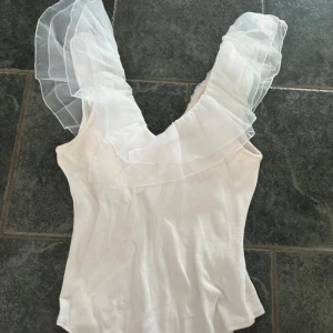 Vit body med volang från Amisu - Supersöt vit body från Amisu med breda, transparenta volangärmar i organza som ger en riktigt drömmig vibe. Toppen har djup v-ringning och är figurnära i modellen. Perfekt för dig som vill sticka ut med en romantisk och trendig look. Den är lite missfärgad på vissa ställen, men det kanske går bort i tvätten.