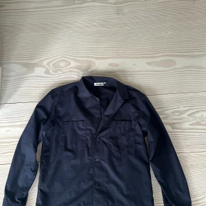 Mörkblå overshirt från weekday - Snygg mörkblå overshirt från weekday med klassisk krage och raka linjer. Skjortan har två bröstfickor, knäppning med tre knappar framtill och långa ärmar med knapp vid manschetten. Materialet är mjuk bomull som känns skönt mot huden. Den är använd en del men är fortfarande i fint skick