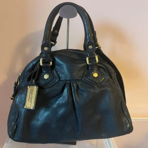 Svart handväska Marc by Marc Jacobs - Cool svart handväska i äkta skinn från Marc by Marc Jacobs med snygga guldfärgade metalldetaljer och dragkedja. Väskan har dubbla handtag, dekorativa sömmar och ett mönstrat innerfoder med logga. Perfekt för dig som gillar stilren design med lyxig känsla.