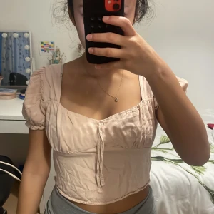 Beige blus från Urban Outfitters - Säljer en beige blus från Urban Outfitters med puffiga korta ärmar och söt knytning framtill. Toppen är croppad och har en mjuk, feminin look med fyrkantig ringning. Materialet känns lätt och har en diskret struktur. Perfekt för dig som gillar romantisk stil.