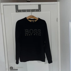 Svart sweatshirt från Hugo Boss - Svart sweatshirt från Hugo Boss med stor BOSS- på bröstet. Tröjan har rund hals, långa ärmar och ribbade muddar. Perfekt för dig som gillar stilrena och klassiska plagg med tydlig branding.