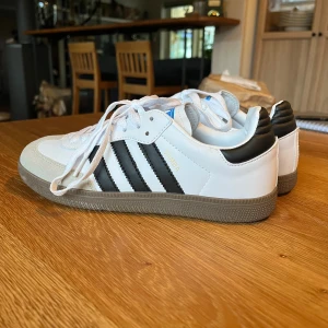 Adidas Samba vita och svarta sneakers - Klassiska Adidas Samba sneakers i vitt och svart med de ikoniska tre ränderna på sidan. Skorna har vit ovandel i skinn, svart häl och detaljer, samt grå mocka vid tån. Gummisula och snörning framtill. Perfekta för en sportig och stilren look.