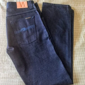 Nudie Organic. Mörkblå dry jeans från Nudie. W31 L32 - Snygga mörkblå dry jeans från Nudie med klassisk femficksdesign, kopparfärgade nitar och knappar samt läderpatch bak. Jeansen har raka ben och är tillverkade i kraftig denim av miljövänlig bomull. Perfekta för dig som gillar stilrena och tidlösa jeans. Dry (otvättad denim). Nytt skick men utan lappar. Motsvarar Grim Tim True Navy i modellen.