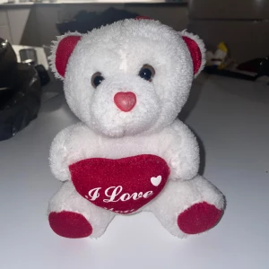 En väldigt gullig nallebjörn  - Detta är en söt vit nallebjörn med röda detaljer och ett hjärta med texten 'I Love'. Perfekt som present eller dekoration. Mjuk och fluffig, i gott skick utan synligt slitage eller defekter. Passar både barn och vuxna som vill ge bort något kärleksfullt.