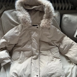 Beige dunjacka med fuskpäls från Shein - Säljer en beige dunjacka från Shein med fluffig fuskpäls runt huvan. Jackan har långa ärmar, två fickor framtill och stängs med både dragkedja och knappar. Perfekt för kalla dagar och har en snygg, neutral look.