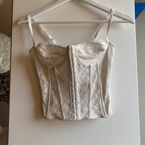 Vit spetskorsett med snörning - Säljer en vit korsett med spetsdetaljer och justerbara axelband. Toppen har snygg snörning i ryggen och hakknäppning framtill. Perfekt för dig som vill ha en trendig och snygg outfit 