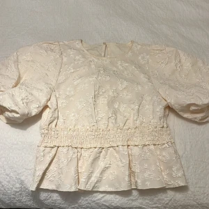 Blommig ljus blus från Dobber - Säljer en ljus blus från Dobber, modell Oksana. Blusen har puffiga korta ärmar, blommigt mönster i strukturen och smockad midja som ger en snygg siluett. Materialet är glansigt och känns lyxigt, med volang nedtill för extra stilpoäng. 