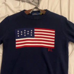 Mörkblå stickad tröja Polo Ralph Lauren - Mörkblå stickad tröja från Polo Ralph Lauren med amerikansk flagga framtill och RL-broderi. Klassisk rund halsringning och långa ärmar. Perfekt för dig som gillar preppy stil och vill sticka ut med en ikonisk detalj.