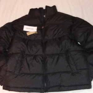 Svart pufferjacka från YOURTURN - Svart pufferjacka från YOURTURN med hög krage och breda, quiltade sektioner. Jackan har dragkedja framtill och en clean, modern look. Perfekt för kalla dagar och enkel att matcha med streetwear. Materialet är syntet och jackan är långärmad.