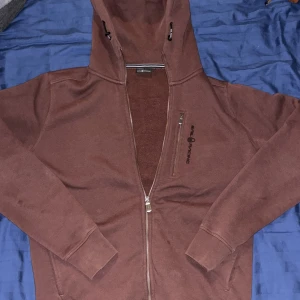 Röd hoodie från Sail Racing med dragkedja - Säljer en röd hoodie från Sail Racing i storlek M. Tröjan har hel dragkedja, huva med snörning och en bröstficka med dragkedja. Mjuk insida och ribbade muddar vid ärmslut och nederkant. Perfekt för chill dagar och snygg streetstil.