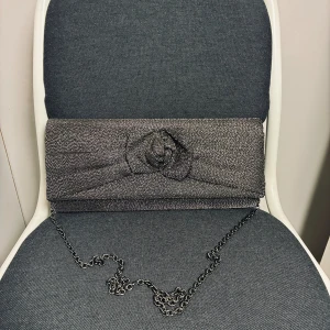 Glittrig silver kuvertväska från Menbur - Snygg kuvertväska från Menbur i glittrigt silver med kedja som axelrem. Framsidan har en dekorativ rosett och insidan är fodrad med svart tyg och har en liten ficka. Perfekt storlek för mobil och småsaker. Stilren och festlig design.