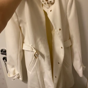 Vit kappa från Atmosphere med bälte - Snygg vit kappa från Atmosphere i storlek 38. Jackan har ett högt krage med dekorativa guldfärgade knappar och ett matchande bälte i midjan med guldfärgat spänne. Fickor framtill och stilren design, perfekt för dig som gillar clean och elegant look. Aldrig använt. Jag är öppen för förslag 