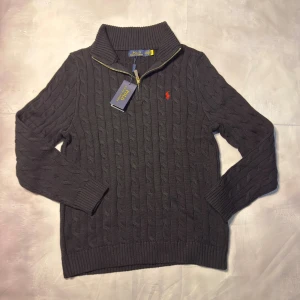 Svart kabelstickad tröja Polo Ralph Lauren - Svart kabelstickad tröja från Polo Ralph Lauren med halv dragkedja och ribbad krage. Tröjan har långärmad design och ett litet rött broderat logomärke på bröstet. Tillverkad i 100% bomull för en mjuk och skön känsla. Perfekt för dig som gillar klassisk stil med en modern twist.