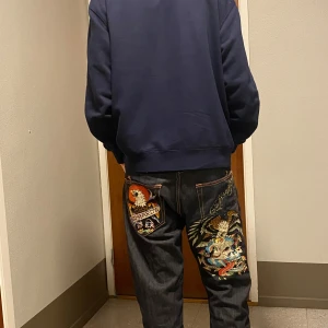 Ed Hardy jeans med broderi, stl 33/34 - Unika mörkblå jeans från Ed Hardy med stora, färgglada broderier på bakfickorna – örn, dödskalle och logga. Klassisk femficksmodell i 100% bomull med raka ben och orangea sömmar. Perfekta för dig som vill sticka ut med coola detaljer.