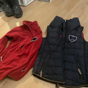 Marinblå dunväst och fleece från Jacson - Marinblå quiltad dunväst från Jacson med emblem på bröstet och dragkedja framtill. Västen har två fickor med dragkedja och en hög krage med mjukt vitt foder på insidan inkl tillhörande fleece . Storlek är xs men passar mer som s 