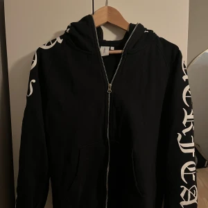Svart hoodie med tryck från Junkyard - Svart hoodie från Junkyard i storlek S med vit text i gotisk stil längs ärmar och över huvan. Tröjan har dragkedja framtill, huva och stora fickor. Perfekt för dig som gillar streetwear och vill sticka ut med coola detaljer. Knappt använd och säljer för är för liten