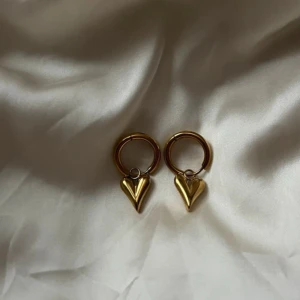 Guldiga örhängen med hjärtanhänge - Snygga örhängen i guldton med rund ring och ett hängande hjärta i metall. Perfekt accessoar för dig som gillar stilrena detaljer med en romantisk touch. Hjärtat är blankt och har en modern, minimalistisk form. Stainless Steel🩷 18k guldpläterad 💞