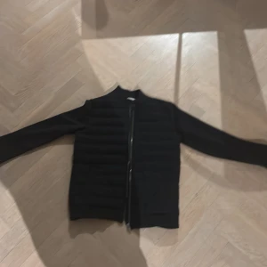 Svart quiltad bomberjacka från M&S - Svart cardigan från M&S med quiltad framsida och insida, hel dragkedja och ribbad krage. Jackan har långärmade släta ärmar och en stilren look som passar perfekt till streetwear. Materialet är syntet och polyester för en lätt och bekväm känsla. Den är knappt använd, som ny skick.