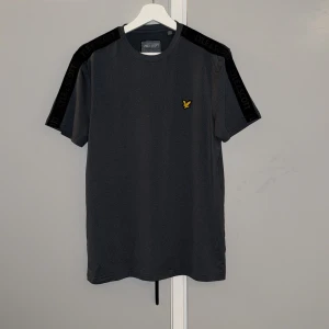 Mörkgrå Lyle & Scott t-shirt med logoband - Snygg mörkgrå t-shirt från Lyle & Scott med svarta logoband längs axlarna och ett gult broderat fågelmärke på bröstet. Rund hals och korta ärmar, perfekt för en clean och sportig look. Materialet är mjuk bomull som känns skönt mot huden.