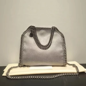 Silver axelväska med kedjedetaljer - Snygg axelväska i silver från Stella McCartney med coola kedjehandtag och kedjekant runt hela väskan. Väskan har en mjuk, glansig yta och en stilren, fyrkantig form. Perfekt för dig som vill sticka ut med en trendig accessoar.