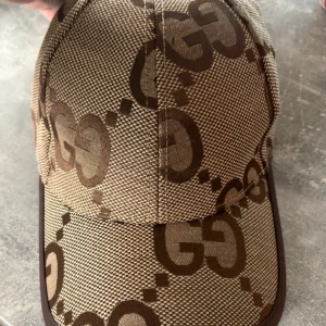 Gucci beige keps med GG-mönster - Snygg beige keps från Gucci med det klassiska GG-logomönstret i brunt. Kepsen har böjd skärm och är tillverkad i ett slitstarkt tyg med bruna detaljer längs kanterna. Perfekt för dig som vill ha en lyxig och trendig accessoar.