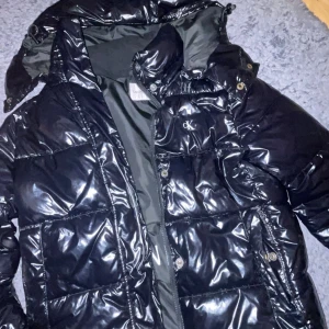 Svart pufferjacka från Calvin Klein - Snygg svart pufferjacka från Calvin Klein Jeans med glansig finish och stor huva. Jackan har dragkedja, tryckknappar och logotyp på bröstet samt insidan. Perfekt för kalla dagar och har en modern, boxig passform.