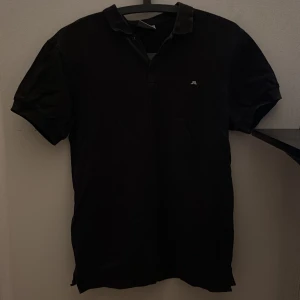 Svart piké från J.Lindeberg, storlek S - Svart pikétröja från J.Lindeberg med klassisk krage och två knappar framtill. Diskret JL-logga broderad på bröstet. T-shirten är kortärmad och tillverkad i mjuk bomull som känns skön mot huden. Perfekt för en clean och stilren look.