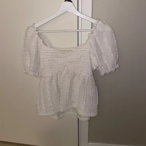 Vit blus med puffärmar - Supersöt vit blus med puffiga korta ärmar och smockad struktur. Blusen har en fyrkantig ringning och volangig nederkant som ger en romantisk vibe. Perfekt för dig som gillar en feminin och trendig stil.